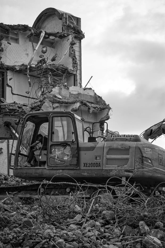 A monochrome image of an excavator demolishing a building in an urban area.